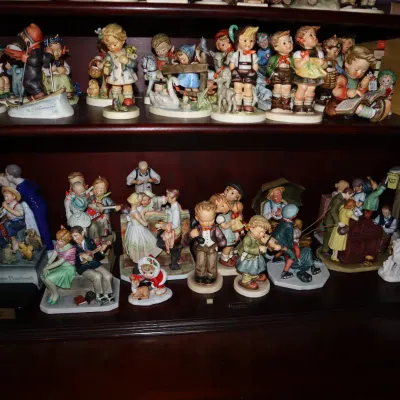 Sale Photo Thumbnail #5: Collection of vintage porcelain figurines, including Hummel-style children and Norman Rockwell characters, displayed on wooden shelves alongside a stack of books.