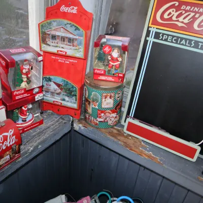 Sale Photo Thumbnail #171: Collection of vintage-style Coca-Cola memorabilia including boxed Santa ornaments, a lunchbox, decorative tin, wooden wall rack, and a chalkboard menu sign.