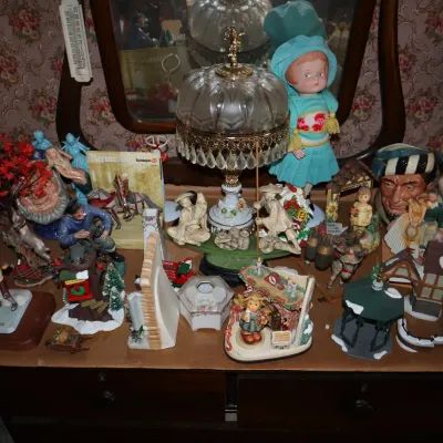 Sale Photo Thumbnail #6: Collection of vintage collectibles including an ornate glass table lamp with crystal prisms, ceramic figurines, Christmas village pieces, and a vintage doll displayed on a wooden dresser.