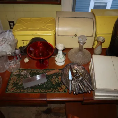Sale Photo Thumbnail #130: Collection of tabletop items including vintage glassware, a red compote dish, silver-plated flatware in a tray, yellow tin bread boxes, and a large art pottery vase.