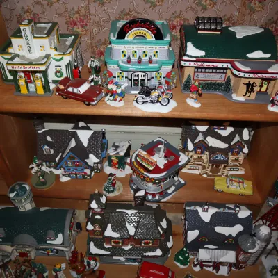Sale Photo Thumbnail #11: Collection of Lemax and Dept 56 lighted Christmas village buildings and accessories displayed on wooden shelves, featuring retro designs including a gas station, record shop, roller rink, and diner.