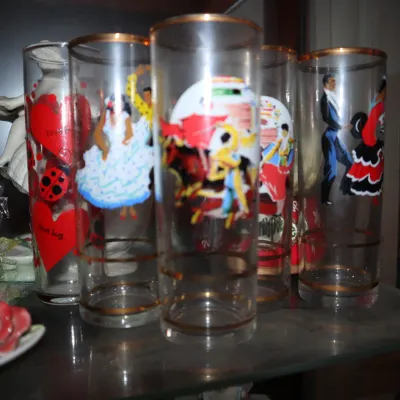 Sale Photo Thumbnail #116: Vintage glass drinking tumblers with gold rims featuring printed Spanish matador, dancer, and ladybug designs, displayed on a cabinet shelf.