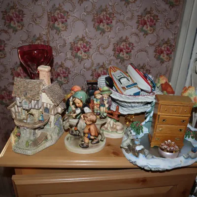 Sale Photo Thumbnail #18: Collection of decorative figurines and knick-knacks, including a cottage structure, Goebel-style figures, a miniature wooden dresser, and small toy boats.
