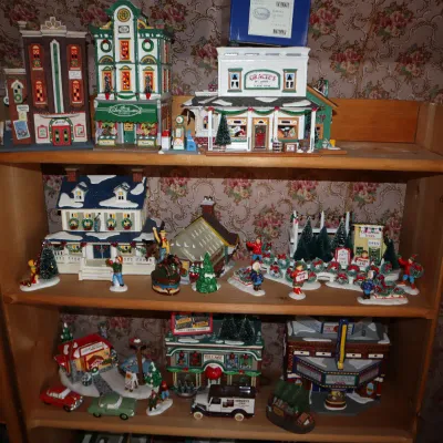 Sale Photo Thumbnail #12: Collection of Christmas village miniature buildings and figurines displayed on wooden shelves, including storefronts, houses, and accessories.