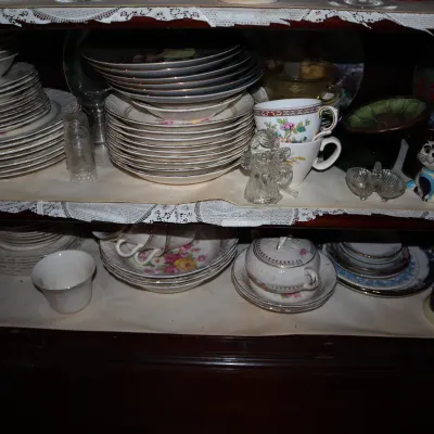 Sale Photo Thumbnail #129: Collection of vintage china and glassware, including stacks of floral-patterned dinnerware, teacups, a clear glass angel figurine, and a small cat figurine displayed on cabinet shelves.