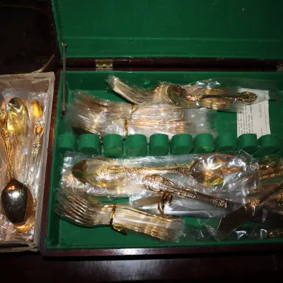Sale Photo Thumbnail #122: Gold-toned flatware set with ornate handles, housed in a green felt-lined wooden storage chest alongside additional pieces in a cardboard box.