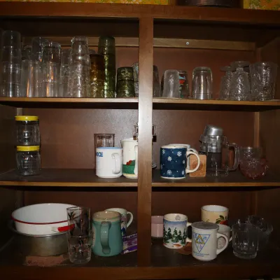 Sale Photo Thumbnail #140: A wooden cabinet filled with vintage glassware, ceramic mugs, and kitchenware, arranged across three shelves.