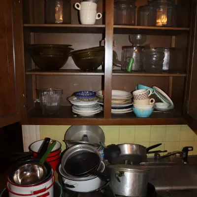 Sale Photo Thumbnail #141: A vintage kitchen filled with glassware, ceramic dishware, and stacked pots and pans, including enamel cookware and glass mixing bowls.