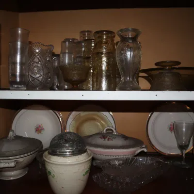 Sale Photo Thumbnail #113: Collection of vintage glassware and dinnerware on wooden shelves, including pressed glass vases, clear tumblers, amber glass, and floral china serving pieces.