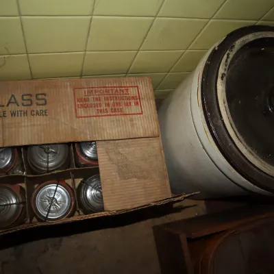 Sale Photo Thumbnail #133: Cardboard box filled with vintage glass canning jars with metal bail closures, sitting beside a large stoneware crock with a lid.