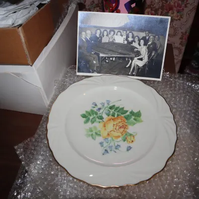 Sale Photo Thumbnail #29: Vintage ceramic dinner plate featuring a central yellow rose floral motif with gold trim along the scalloped rim, displayed alongside a black-and-white group photo.