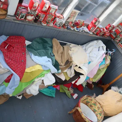 Sale Photo Thumbnail #169: A large pile of vintage clothing laid out along a porch bench, featuring a red tweed jacket and colorful knit sweaters, with a shelf of Coca-Cola collectibles lined above.