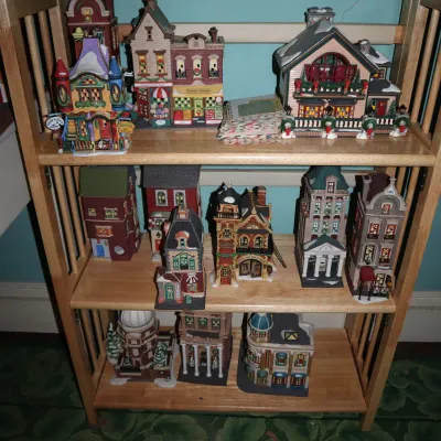 Sale Photo Thumbnail #62: Collection of Dept 56 lighted Christmas village building figures arranged on a three-tier light wood bookshelf.