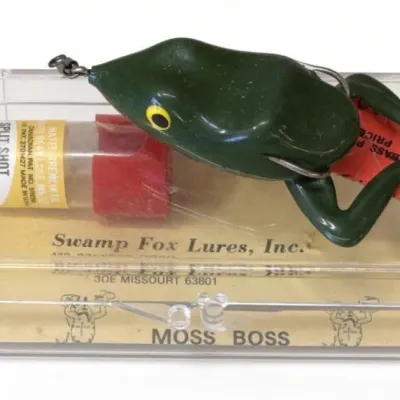 Sale Photo Thumbnail #110: Lot 183: Vntg Swamp Fox Lures “Moss Boss” frog-style fishing lure in clear plastic case. Green soft-plastic frog with yellow eye and hook, resting on the case. Case labeled Swamp Fox Lures, Inc. (Missouri)