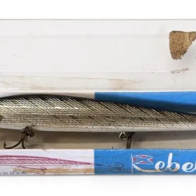 Sale Photo Thumbnail #125: Lot 198: Rebel fishing lure in original blue-and-white cardboard box with clear plastic lid. Long silver/gold scale-pattern hard bait with yellow eye and treble hooks. Box marked “Made in U.S.A.” and “Rebel.” Classic vintage Rebel lure