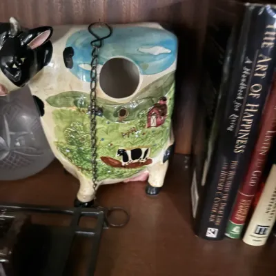 Sale Photo Thumbnail #14: Novelty ceramic cow-shaped birdhouse displayed on a wooden shelf next to books.