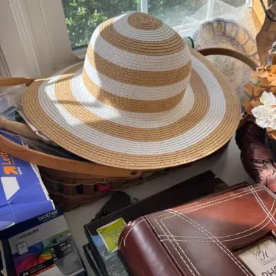 Sale Photo Thumbnail #219: Wide-brimmed woven straw sun hat with alternating tan and white stripes, resting inside a wooden basket near a window sill.