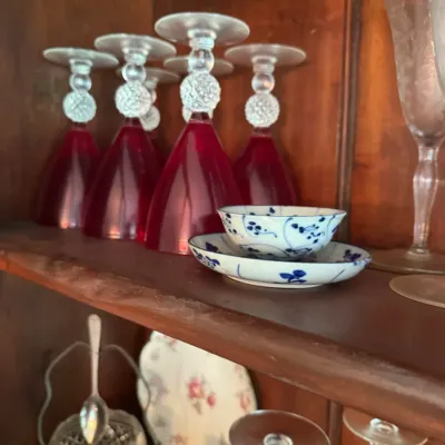 Sale Photo Thumbnail #391: Set of inverted red glass wine goblets with clear textured stems alongside a small blue and white porcelain teacup and saucer inside a wooden cabinet.