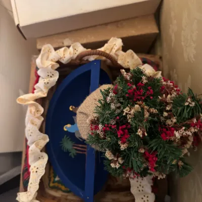 Sale Photo Thumbnail #355: A collection of holiday decor items inside a eyelet-trimmed basket, including a miniature dried flower tree and a blue hand-painted Nativity shelf piece.