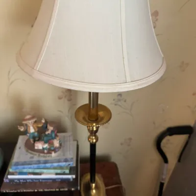 Sale Photo Thumbnail #162: Traditional brass and black table lamp with a tall cream bell shade, resting on a wooden side table alongside small decorative figurines.