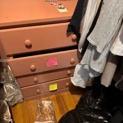 Sale Photo Thumbnail #150: Dusty rose painted wooden dresser with five drawers, set inside a closet alongside hanging garments.