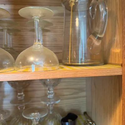 Sale Photo Thumbnail #240: Vintage clear glass beverage pitcher with gold-gilt trim shown alongside upside-down glassware on wooden cabinet shelves.
