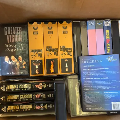 Sale Photo Thumbnail #352: Cardboard box packed with vintage VHS tapes and DVDs, including The Godfather trilogy box set, Johnny Carson collections, Home Alone, and Microsoft Office training video.