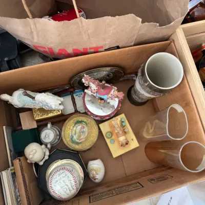 Sale Photo Thumbnail #217: Cardboard box filled with assorted knick-knacks and collectibles, including carousel horse figurines, glassware, porcelain trinkets, and an autographed baseball.