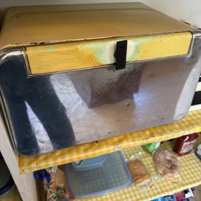 Sale Photo Thumbnail #248: Vintage metal and chrome bread box showing significant wear, paint chipping, and discoloration, sitting on a pantry shelf lined with yellow gingham paper.