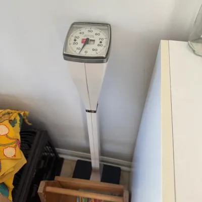Sale Photo Thumbnail #214: Vintage white standing doctor's scale with a large analog dial gauge, positioned next to a small wooden magazine holder.