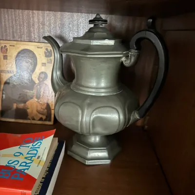 Sale Photo Thumbnail #113: Antique pewter coffee pot with an octagonal pedestal base, paneled body, and dark wooden handle, displayed on a wooden shelf beside books and a small religious icon.
