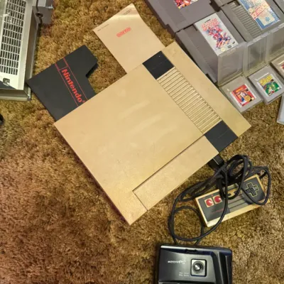 Sale Photo Thumbnail #9: Vintage Nintendo Entertainment System (NES) console with controller, game cartridges, and a Lavec Supervision 35mm point-and-shoot camera on carpet. The NES console exhibits noticeable yellowing to the plastic shell.