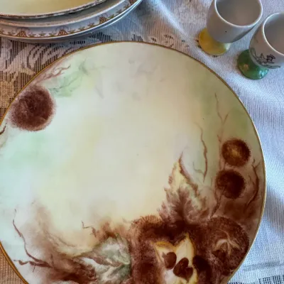 Sale Photo Thumbnail #422: Hand-painted porcelain charger plate featuring a botanical motif in earth tones, shown alongside additional vintage dinnerware and egg cups.