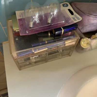 Sale Photo Thumbnail #155: Clear acrylic makeup organizer filled with various cosmetics, including Estée Lauder items, topped with a pack of GE light bulbs.