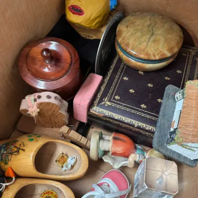 Sale Photo Thumbnail #134: Cardboard box filled with assorted knick-knacks including souvenir wooden clogs, a leather-bound keepsake box, an alabaster trinket dish, figurine, and handmade crafts.
