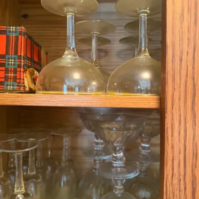 Sale Photo Thumbnail #239: Collection of clear glassware, including stemware and coupe glasses, arranged on wooden cabinet shelves lined with yellow plaid contact paper.