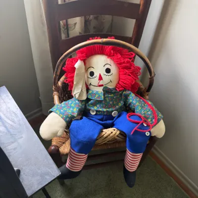 Sale Photo Thumbnail #106: Vintage Raggedy Andy cloth doll with red yarn hair, floral shirt, and blue overalls, seated in a small wicker chair.