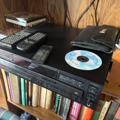 Sale Photo Thumbnail #42: Sony 5-disc CD changer player with three remote controls and a leatherette CD binder sitting on a shelf.