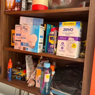 Sale Photo Thumbnail #210: Open wooden wall cabinet filled with household utility supplies, including LED lightbulb packs, Zevo insect trap refills, WD-40, and various small cleaning and repair items.