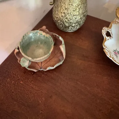 Sale Photo Thumbnail #60: Handmade studio pottery cup resting on a leaf-shaped saucer, alongside an embossed brass pitcher on a wooden surface.