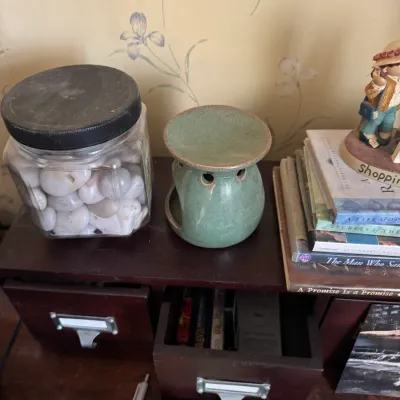 Sale Photo Thumbnail #163: Collection of tabletop decor including a glass jar filled with polished white stones, a ceramic essential oil burner, books, and a figurine set atop a small wooden drawer unit.
