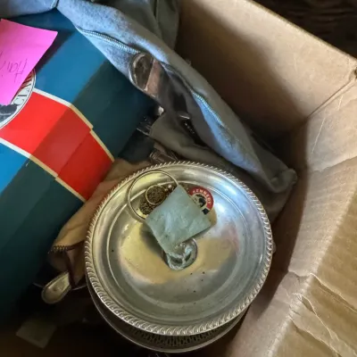 Sale Photo Thumbnail #360: Cardboard box containing a silver-tone dish with a vintage Kennedy campaign pin, silver storage pouches, and miscellaneous household items.