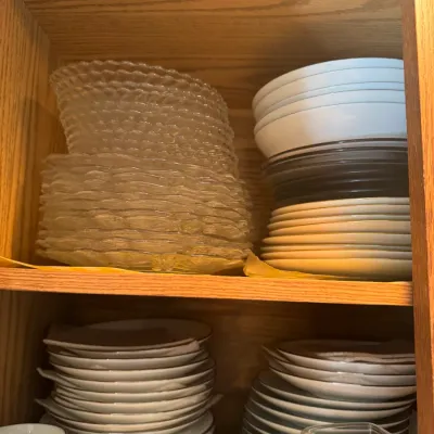 Sale Photo Thumbnail #231: Collection of assorted dinnerware and glassware, including stacks of clear textured glass plates, ceramic bowls, silver-rimmed dishes, and a ceramic cream pitcher, stored inside a wooden cabinet.