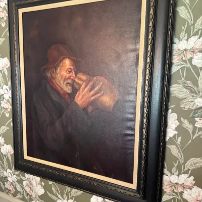 Sale Photo Thumbnail #59: Framed oil painting depicting an elderly bearded man drinking from a jug, signed by the artist at the bottom right. Housed in an ornate black wood frame with gold filigree details and a cream linen liner.