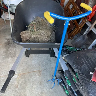 Sale Photo Thumbnail #203: Black poly wheelbarrow with metal handles, shown alongside a blue and yellow rotary garden cultivator and bags of mulch.