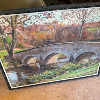 Sale Photo Thumbnail #330: Framed impressionist oil painting depicting a stone arch bridge over a river set against an autumn landscape, presented in a dark carved wooden frame.