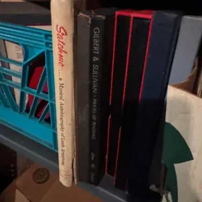Sale Photo Thumbnail #85: A collection of vintage vinyl record sets and music books stored on a shelf, including Louis Armstrong's 'Satchmo' autobiography and Gilbert & Sullivan's 'Pirates of Penzance'.