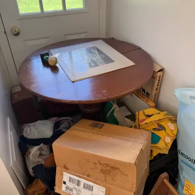 Sale Photo Thumbnail #215: Round wooden drop-leaf pedestal table holding matted artwork, surrounded by various household items and boxes.