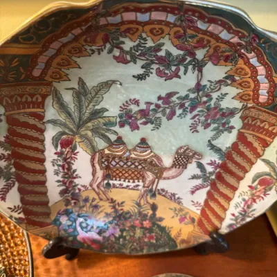 Sale Photo Thumbnail #50: Decorative hand-painted porcelain platter featuring a Middle Eastern scene with a camel, palm tree, and ornate columns in vibrant colors.