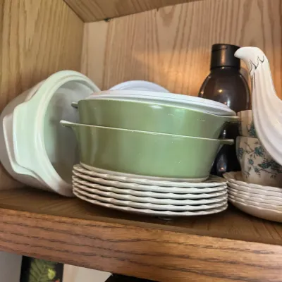 Sale Photo Thumbnail #269: Collection of vintage kitchenware stored in a wooden cabinet, including avocado green Pyrex-style casserole dishes, stacked scalloped plates, and floral teacups.
